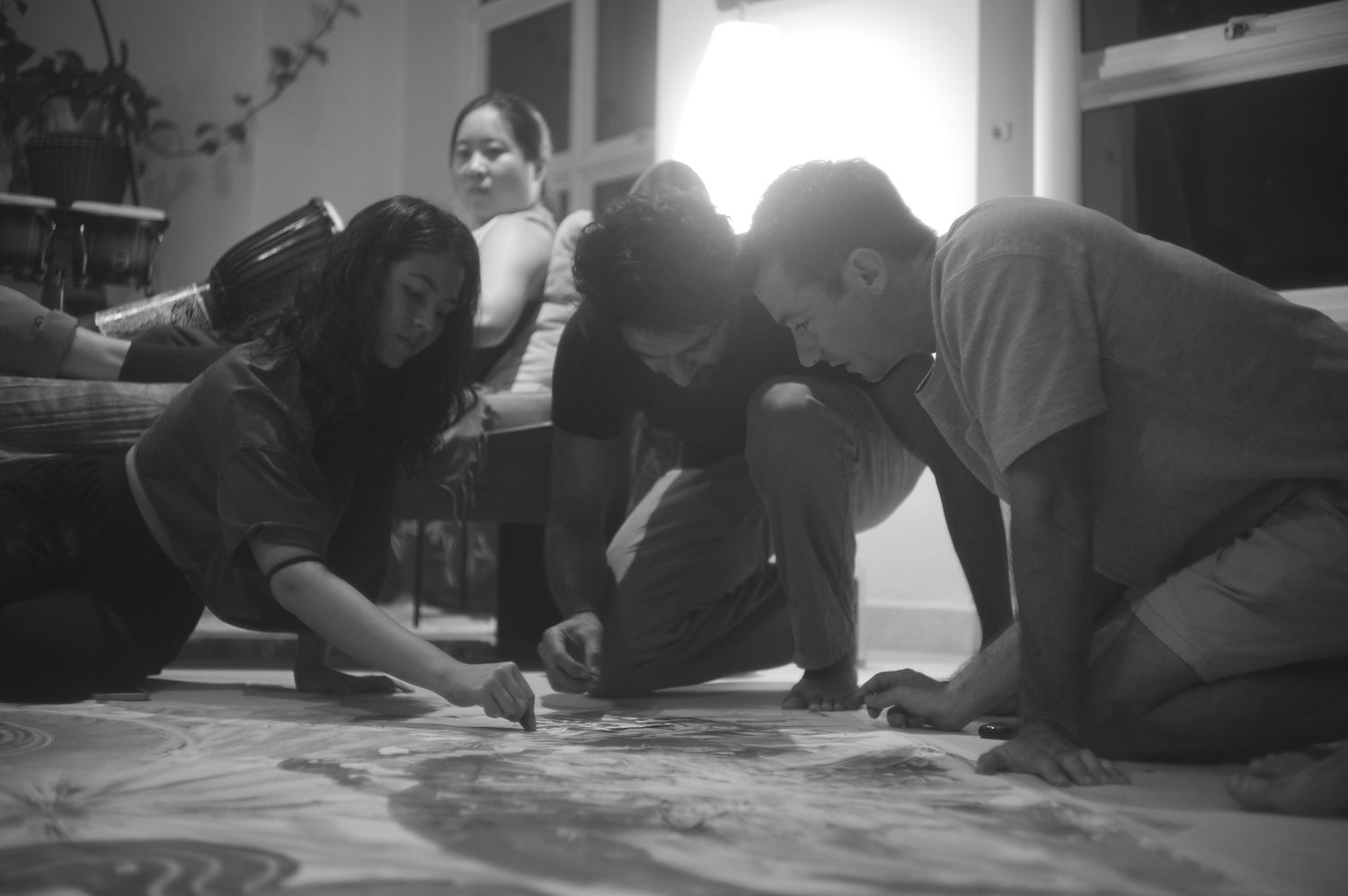 Four people kneeling around a canvas on the floor, working together by lamplight